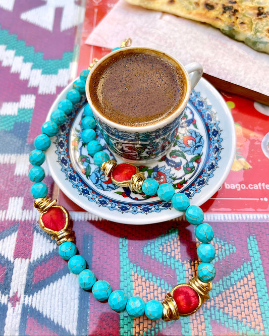 A turquoise beaded necklace with red and gold accents is draped around a small cup of Turkish coffee on a patterned saucer, with a piece of flatbread in the background.