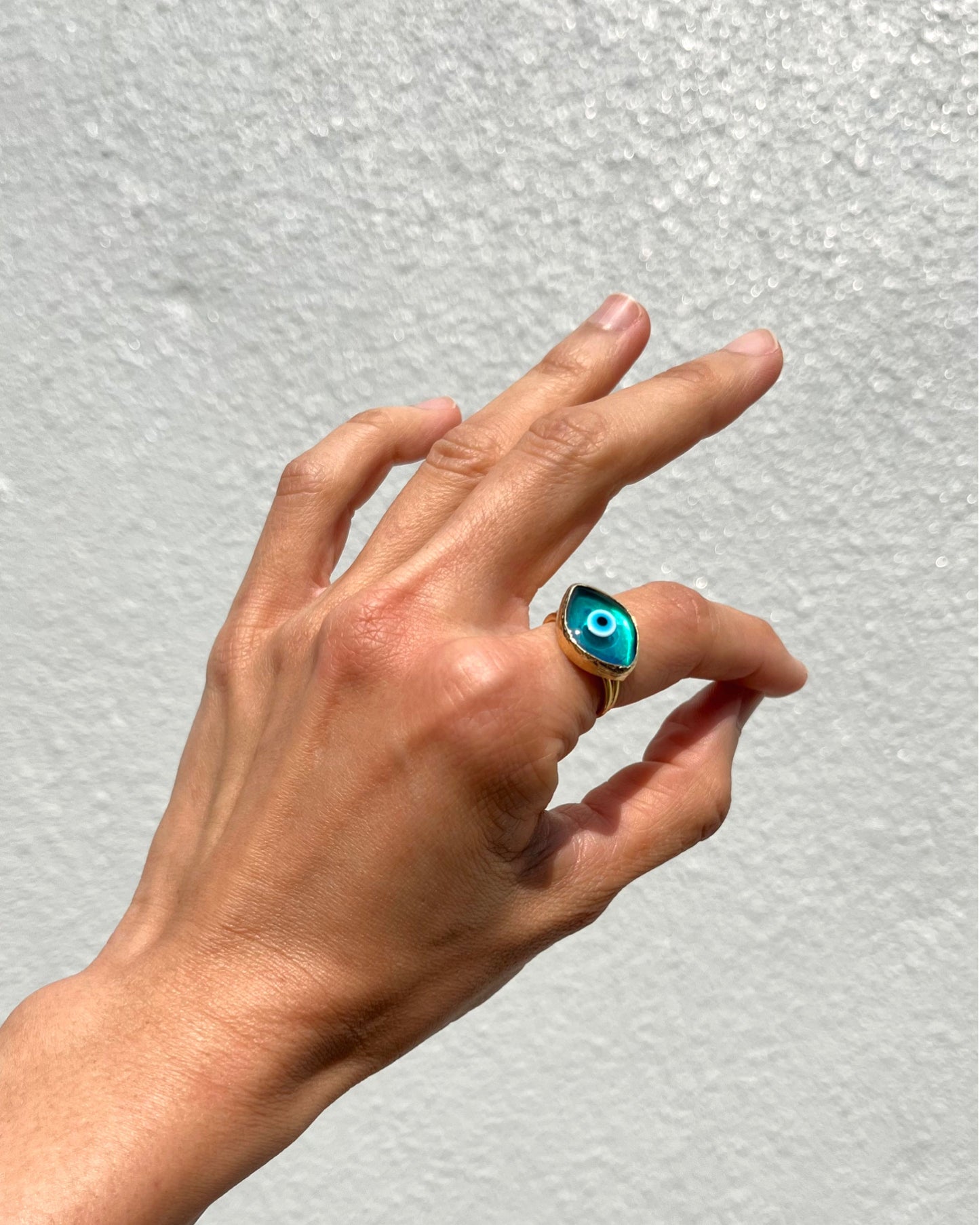 A gold ring with a turquoise blue evil eye stone, worn on a person's ring finger against a textured white wall.