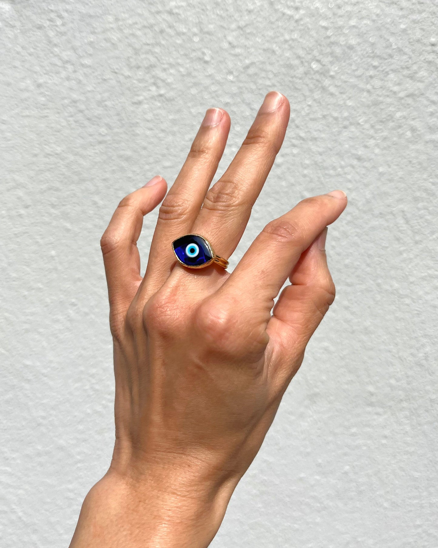 A hand wearing a ring with a blue and white evil eye design on a metallic band, set against a textured light gray wall.