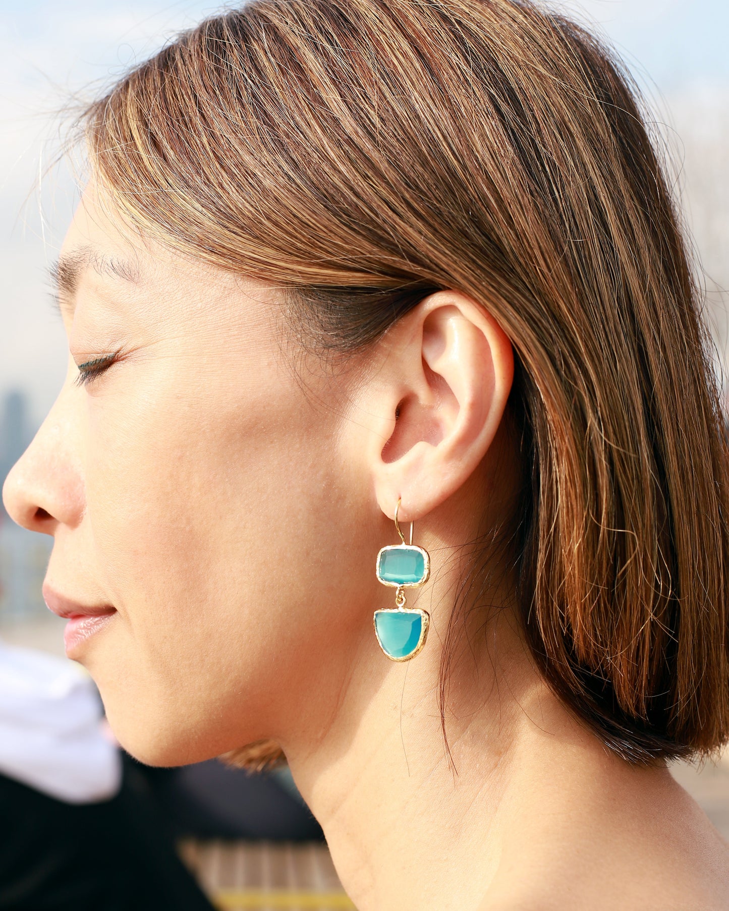 A woman in profile wearing a pair of gold drop earrings with two stacked turquoise-blue stone charms, photographed outdoors in natural light.